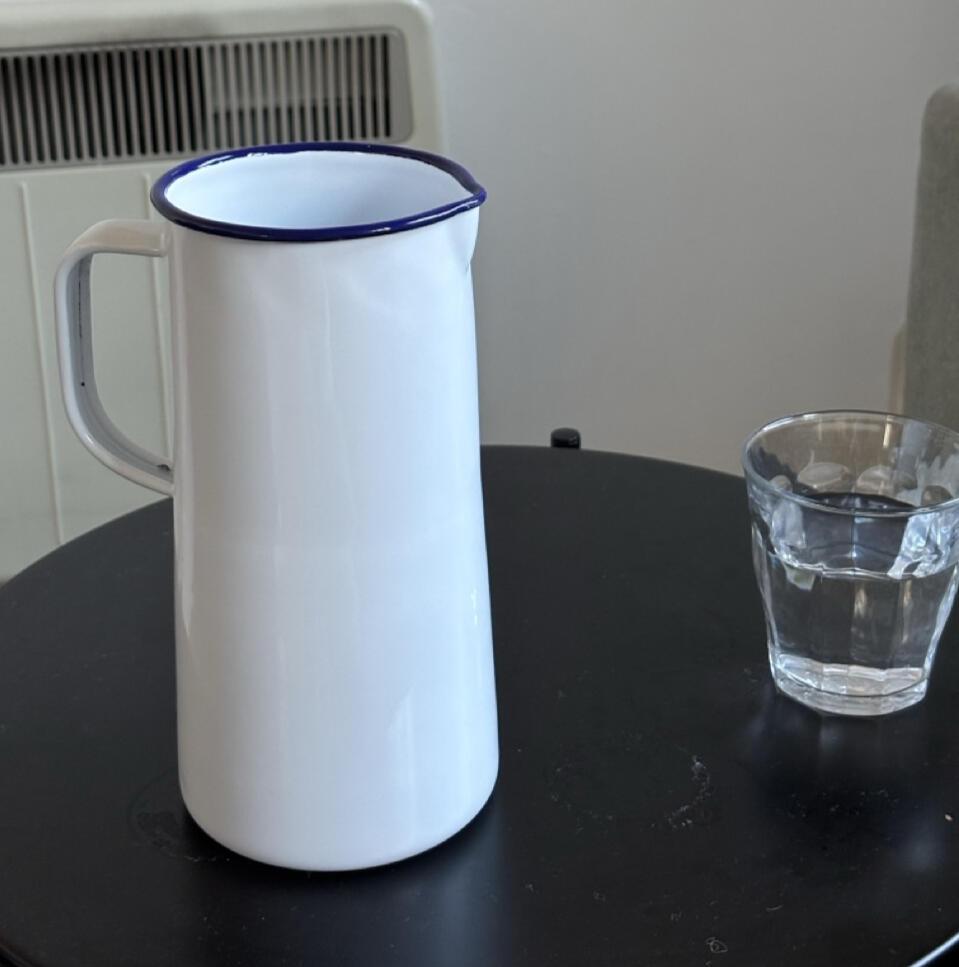 A white enamelware pitcher with a blue rim, sitting on a black metal table with a glass of water next to it