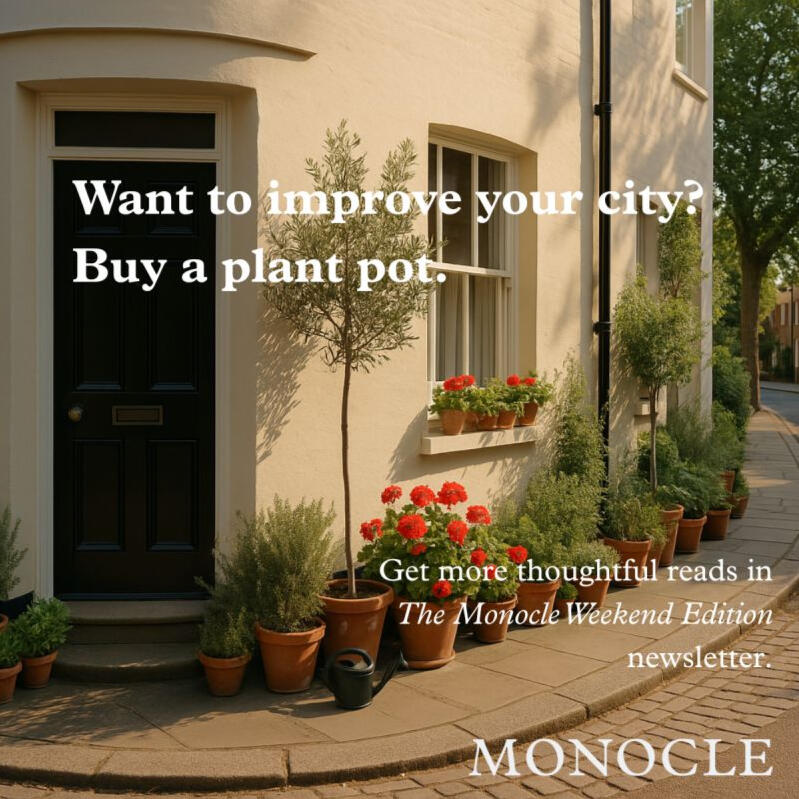 A photo of a quaint city street that wouldn't look out of place somewhere in a leafy suburb of a nondescript European city. On the footpath, up against the exterior wall of a house, there are in excess of 10 pots with various plants and flowers growing in