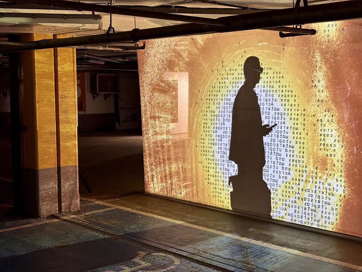 A photo taken inside an indoor carpark. A screen is being hung from the ceiling with an orange projection shone onto it. A shadow of a person can be seen, as though they are walking between the projector and the screen.