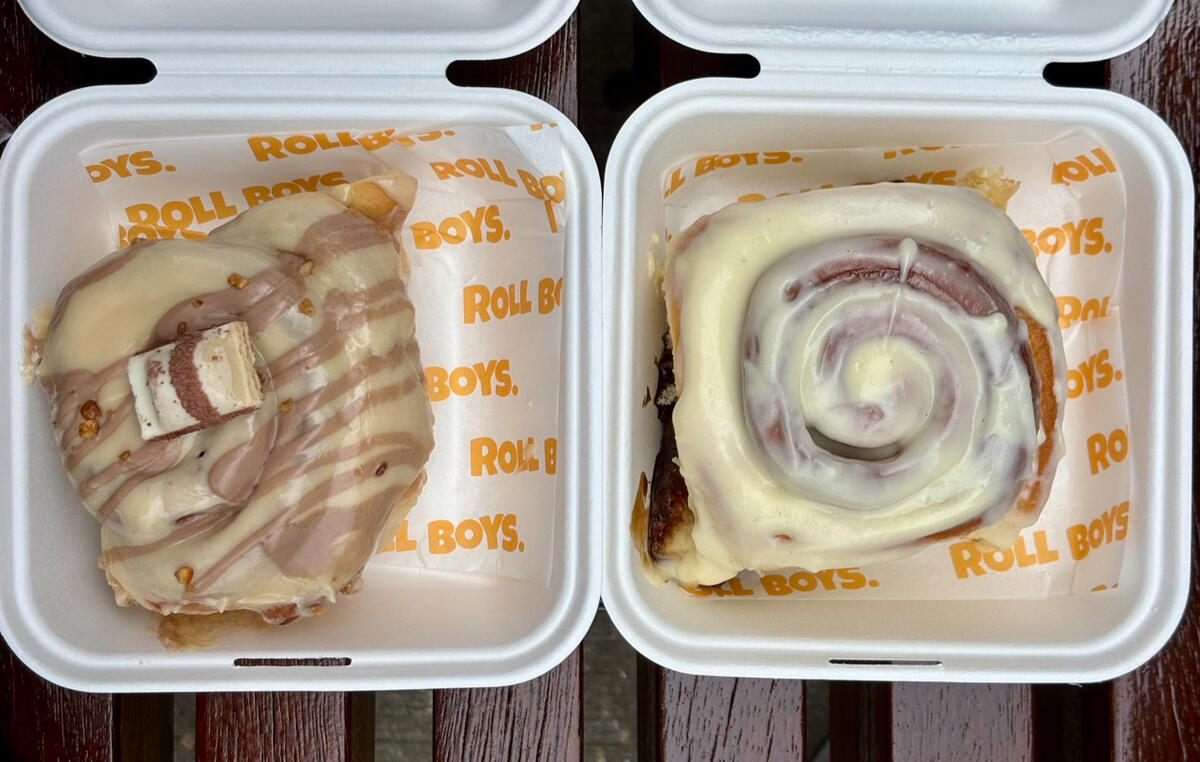 A topdown photo of two cardboard takeaway boxes, each with a cinnamon bun in it. They are liberally coated in frosting. The one on the left is brown and off-white, with a square of Kinder Bueno chocolate. The one on the right is coated in a more yellowy-wh